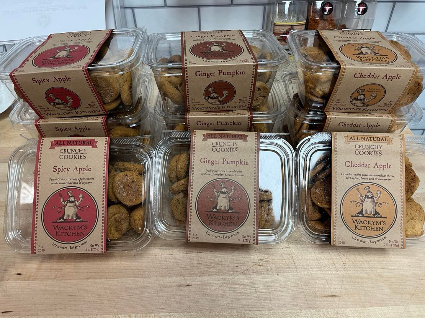 Six containers of cookies with brown labels, varying flavors like