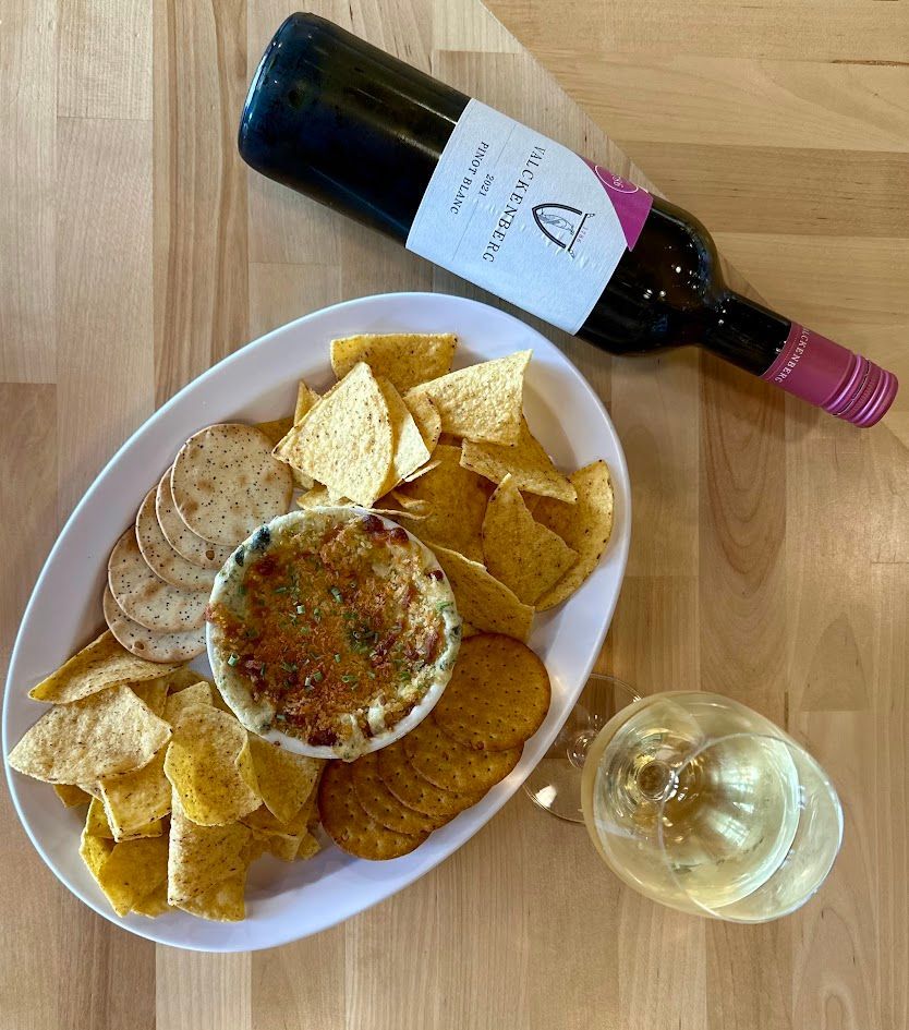 A white platter with dip, chips, crackers, a wine bottle, and a wine glass on a wood table.