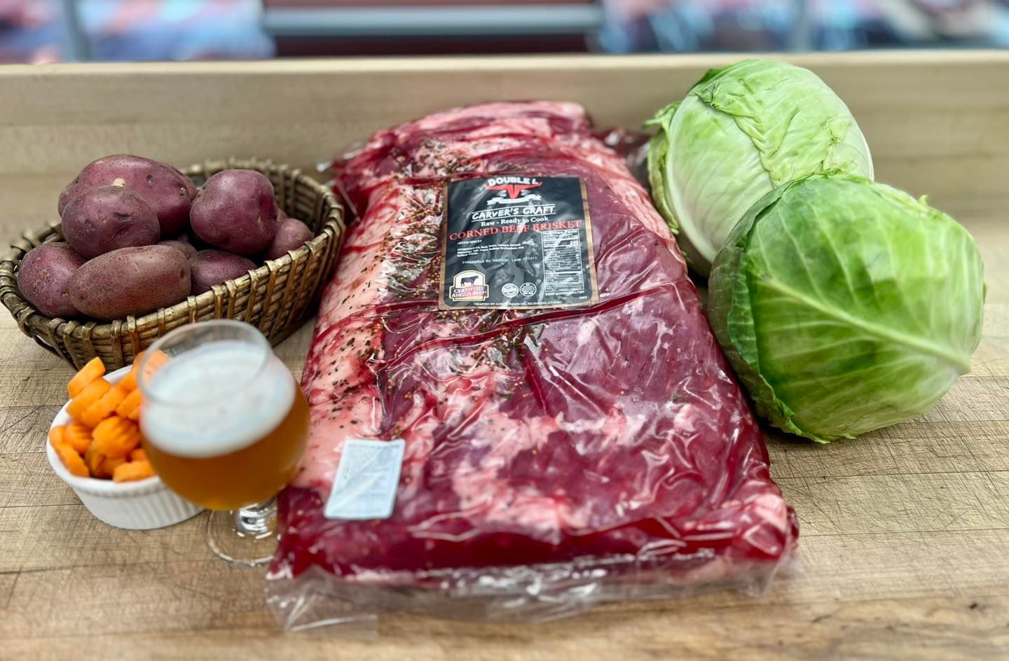 A raw beef brisket, cabbage, potatoes, carrots, and a beer on a butcher block.