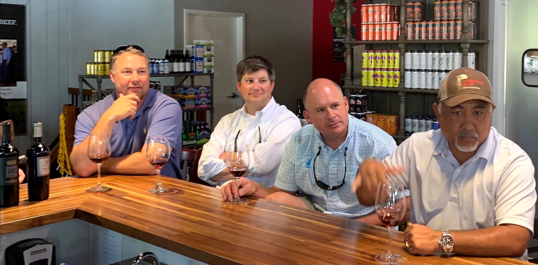 Four men seated at a bar, holding wine glasses. They are in a wine shop setting, with shelves in the background.