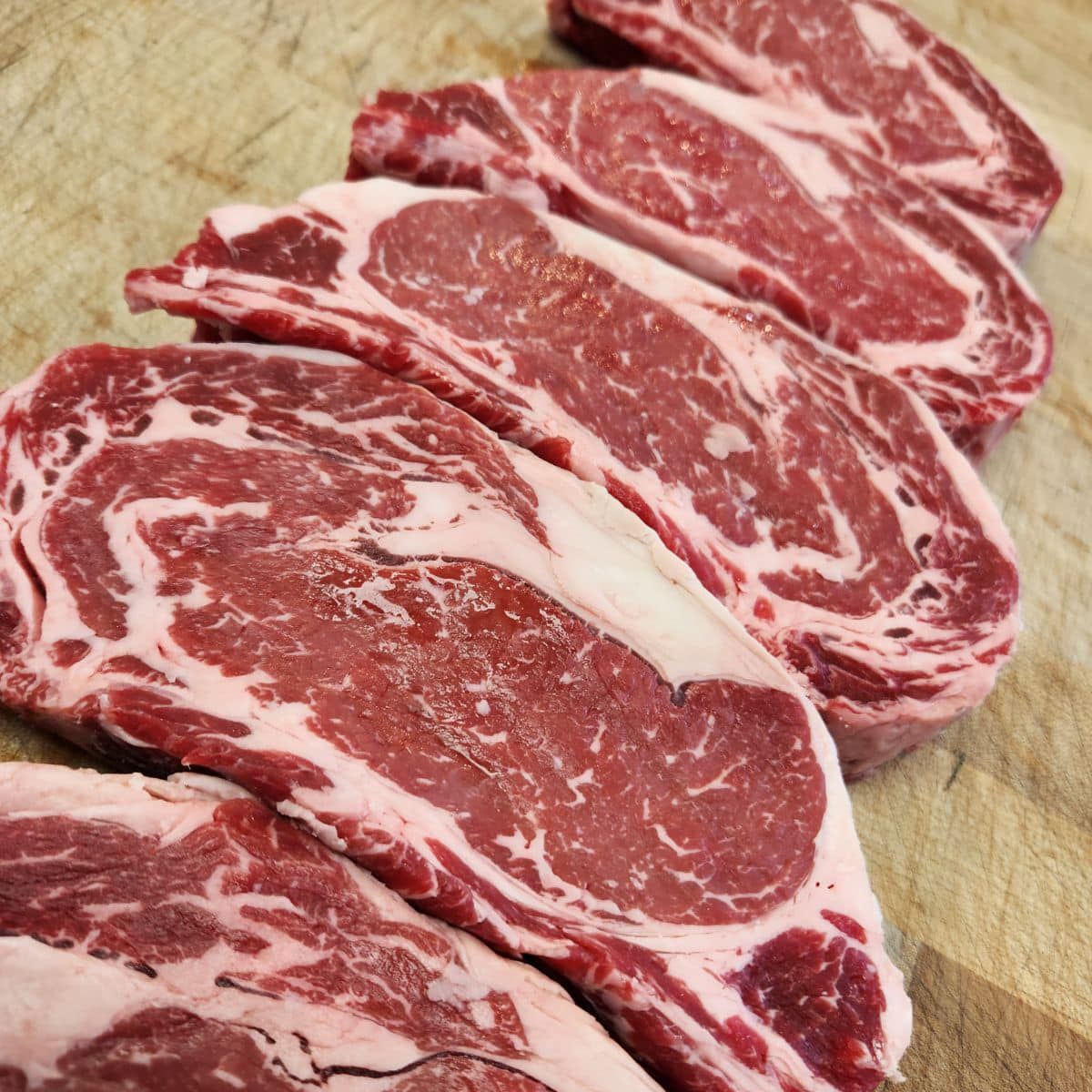 Raw ribeye steaks arranged diagonally on a wooden surface, showing marbling and red color.