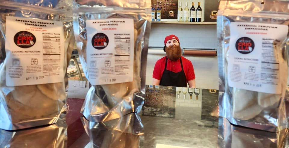 A man with a beard in a red shirt stands behind a counter, with bags of food in front of him.