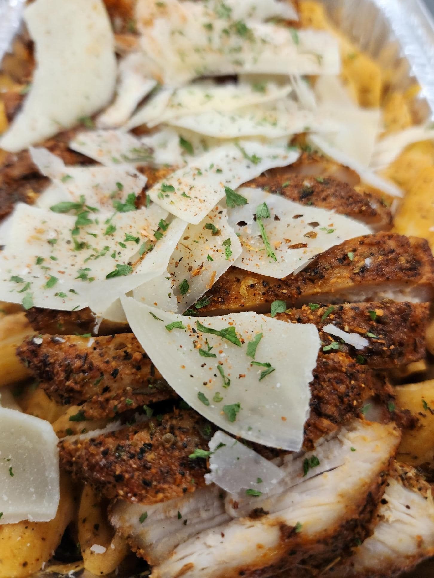 Chicken with shaved Parmesan and herbs on a bed of yellow food in a foil container.
