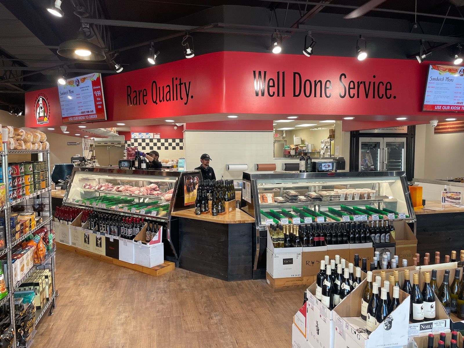 Interior of a butcher shop with display cases, red signage, and a worker behind the counter.