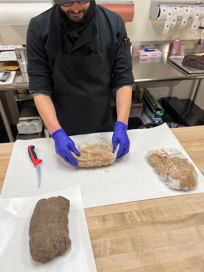 Chef in black uniform, preparing meat on butcher paper. Blue gloves, smiling. Two cooked meat slabs on table.