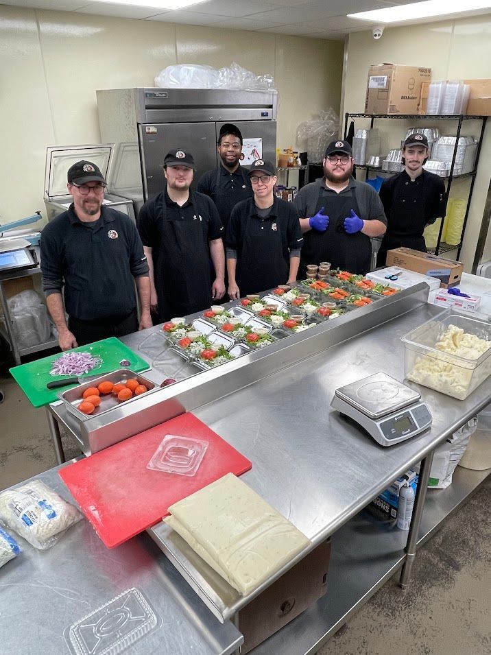 Restaurant staff prepares food at a stainless steel counter. Tomatoes, vegetables, and ingredients are displayed; staff members smile.