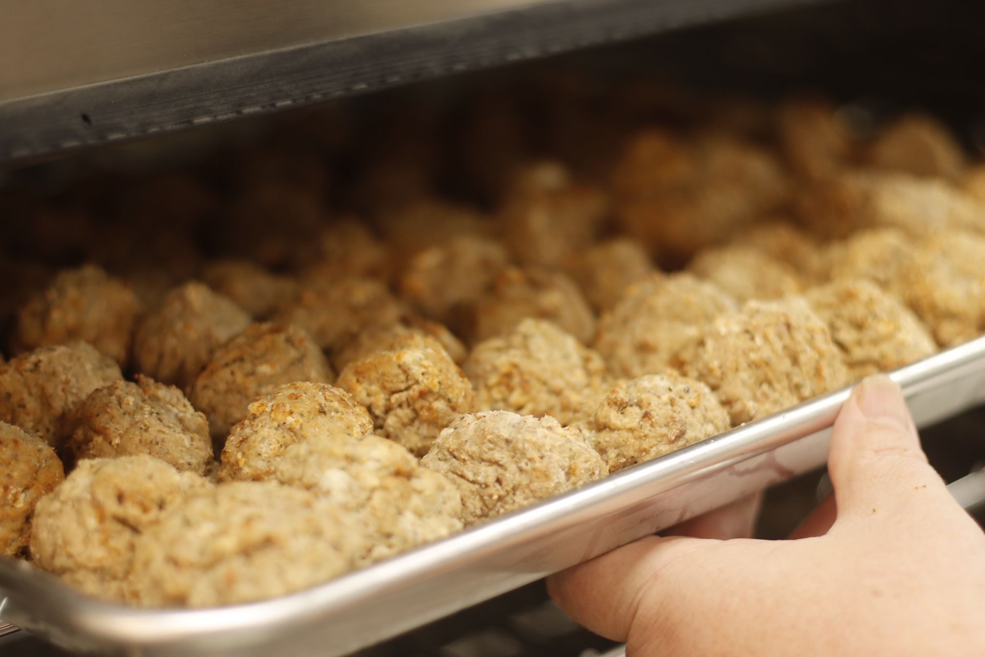 Hand holding a tray of baked meatballs, inside a warm oven.