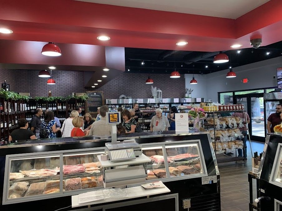 Inside a shop, a crowd gathers around a counter with displayed meats, red ceiling, and overhead lights.