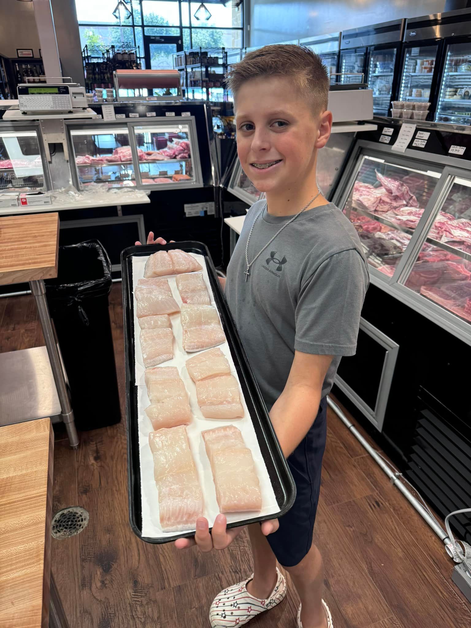 Young boy holding a tray of raw fish fillets, smiling, in a butcher shop.