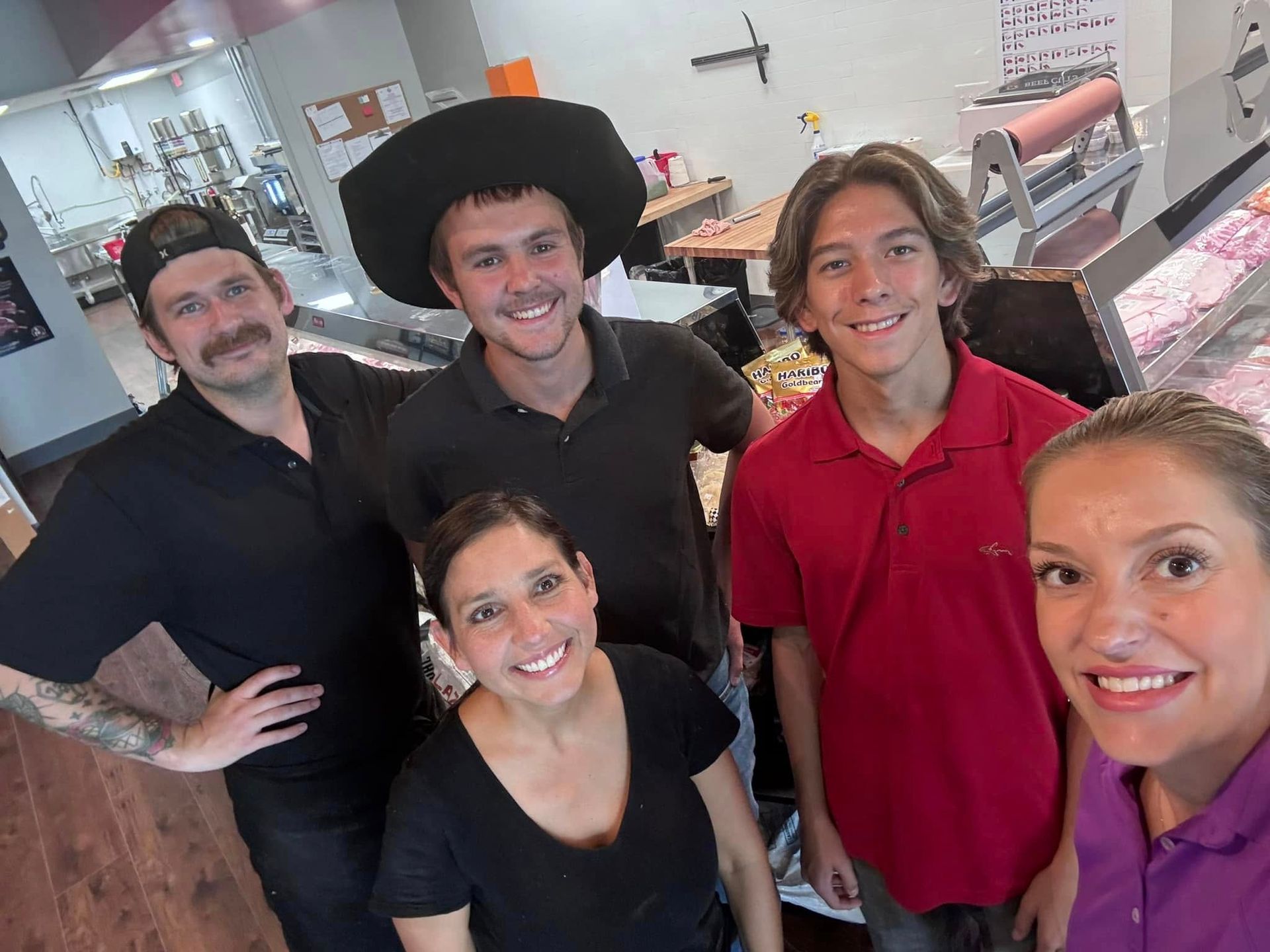 Five people smiling inside a deli. Various skin tones, dark-colored shirts. Behind is a counter with food.