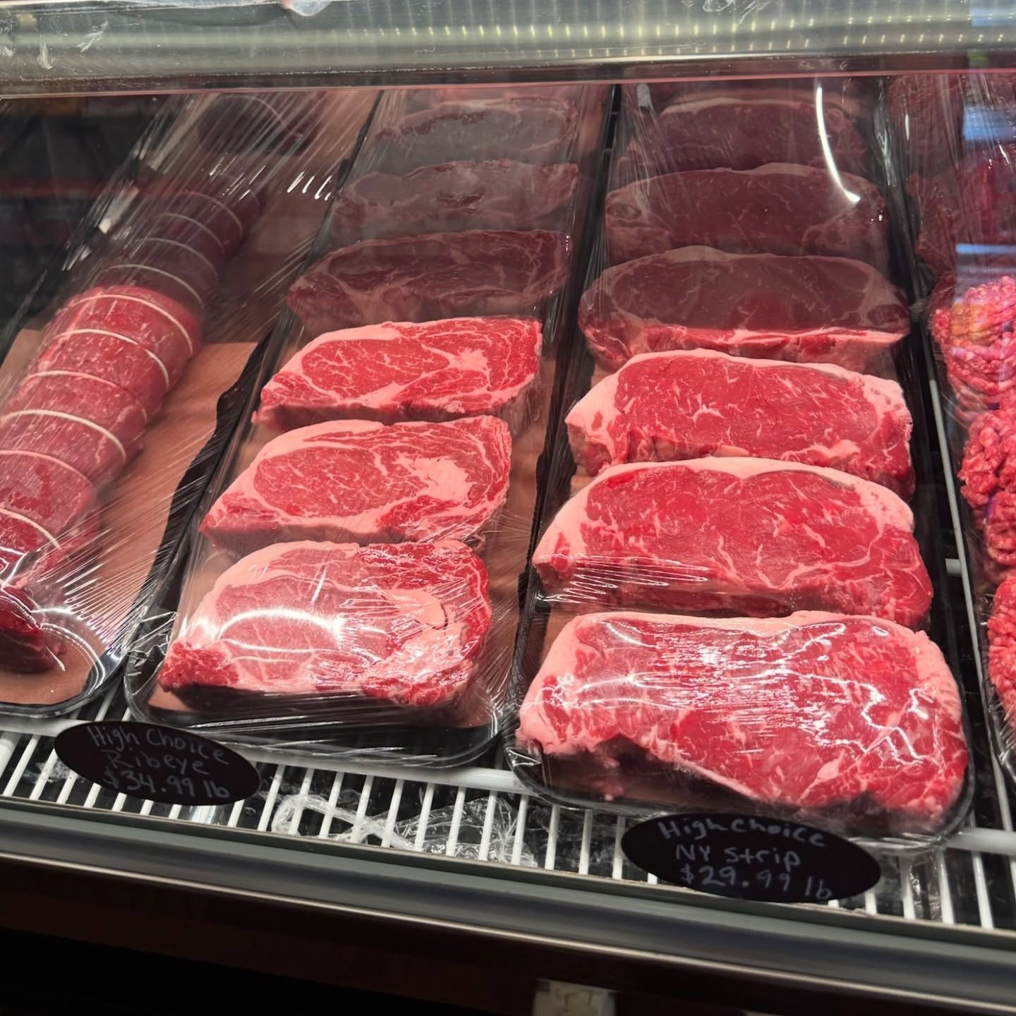 Butcher shop display case with various cuts of red meat, including steaks and a tied roast.
