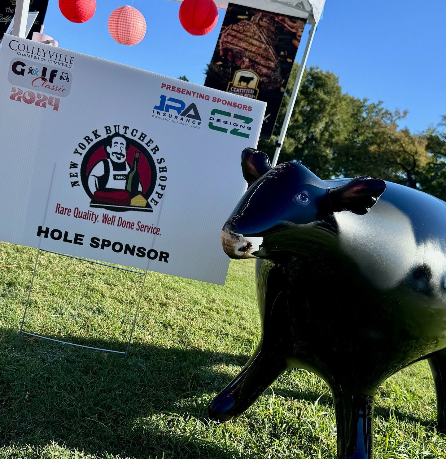 Black cow statue beside a sign for New York Butcher Shoppe as a hole sponsor at a golf event.