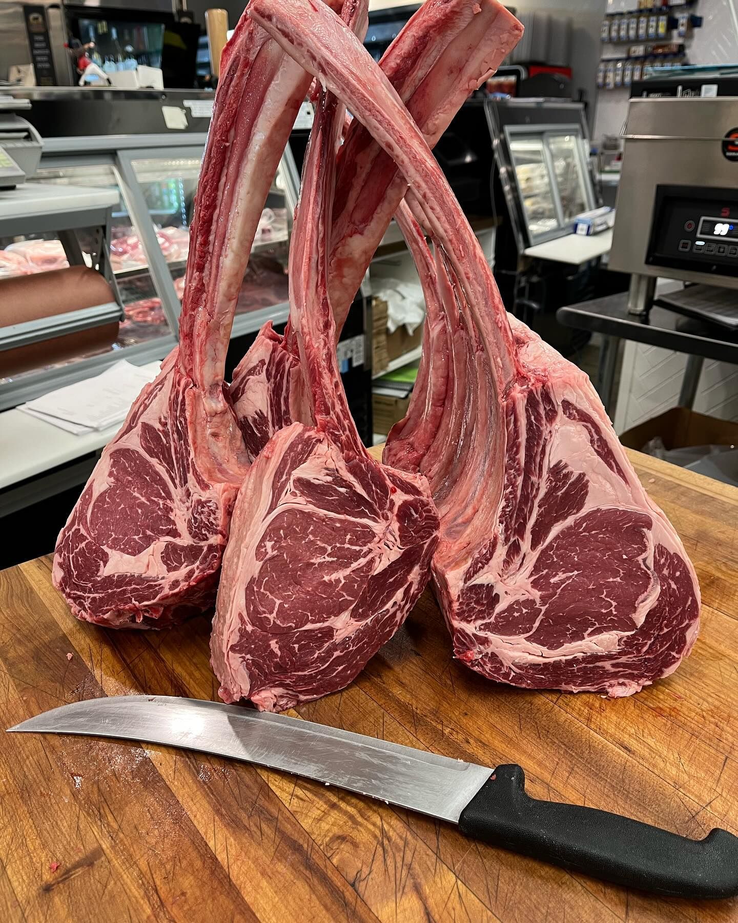 Three raw tomahawk steaks and a knife on a wooden cutting board in a butcher shop.