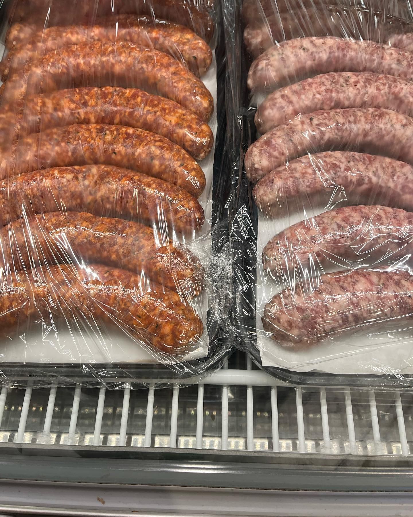 Two trays of sausages, one reddish-brown and one pink, wrapped in plastic in a refrigerated display case.