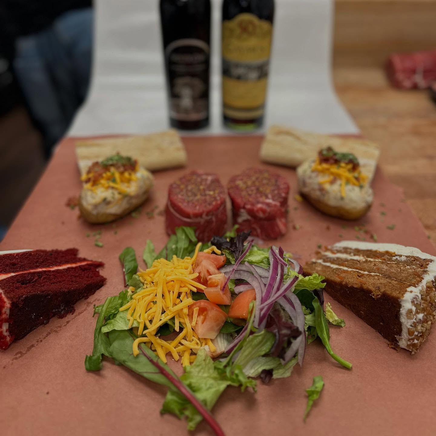 Steaks, baked potatoes, salad, cake, and wine bottles on a pink surface.