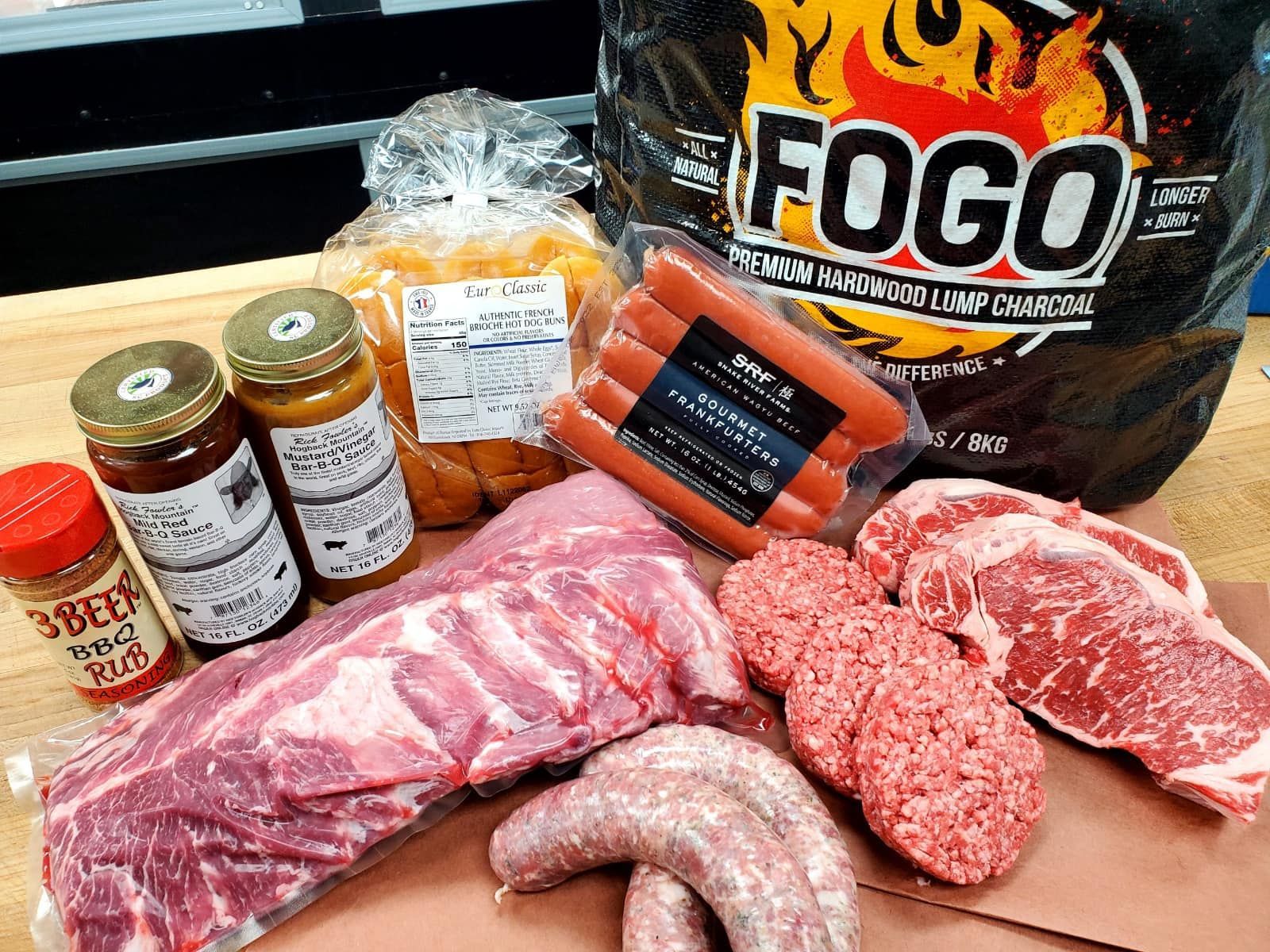 Assorted meats and condiments, including ribs, steaks, sausages, hotdogs, and Fogo charcoal, ready for grilling.