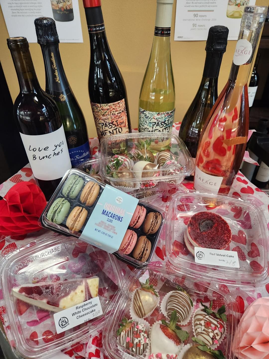 Valentine's Day treats: wine bottles, macarons, cheesecake, chocolate-covered strawberries on a table with hearts.