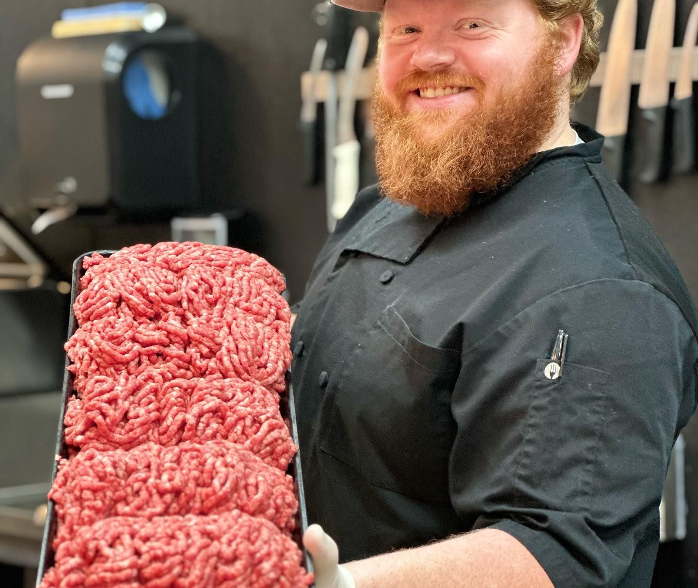 Butcher holds tray of ground meat, smiling. Wearing black chef's coat, hat, beard; knives and equipment in background.