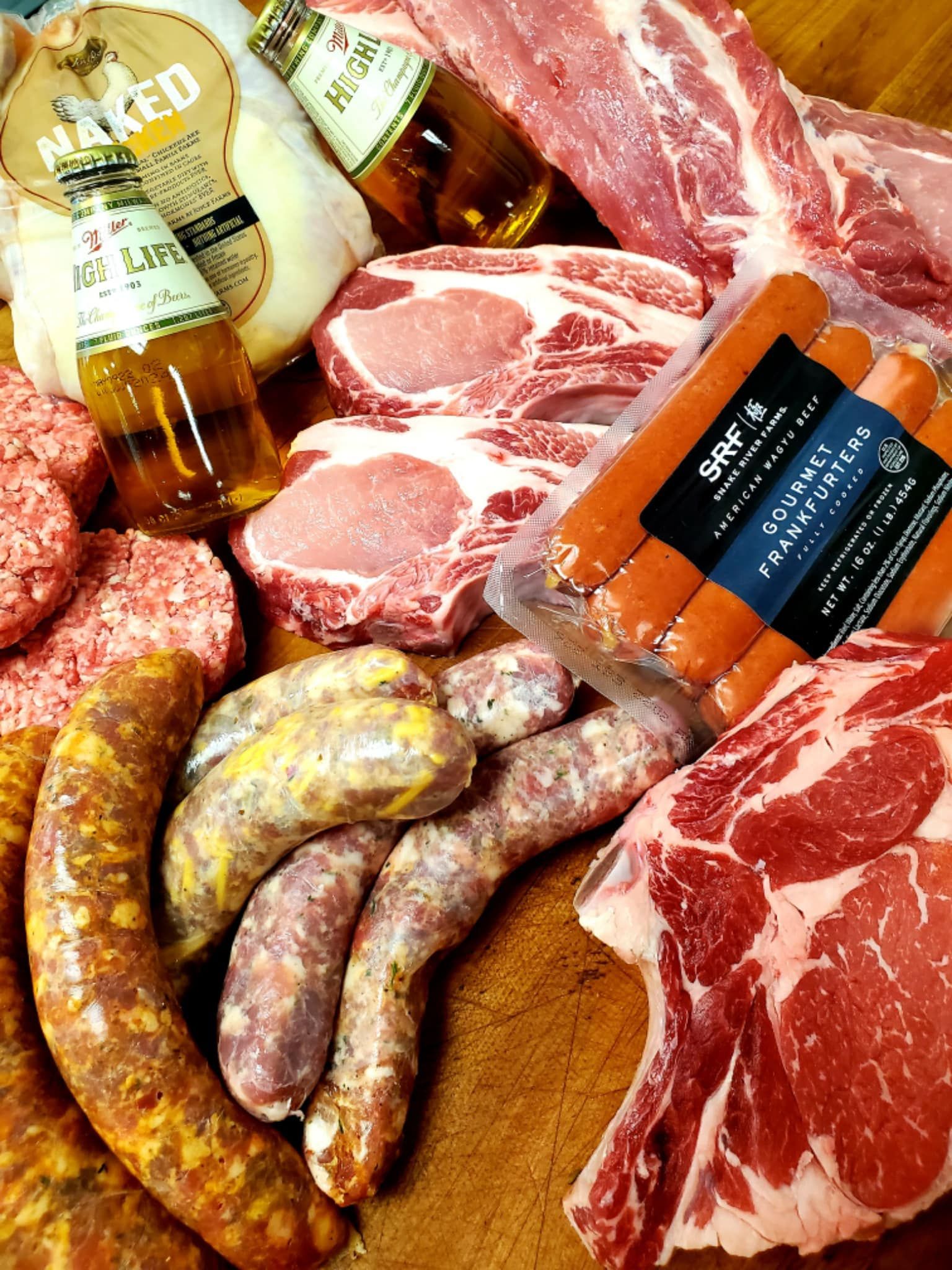 Assortment of raw meats: sausages, pork chops, ground beef, hot dogs, and a large steak, with beer bottles.