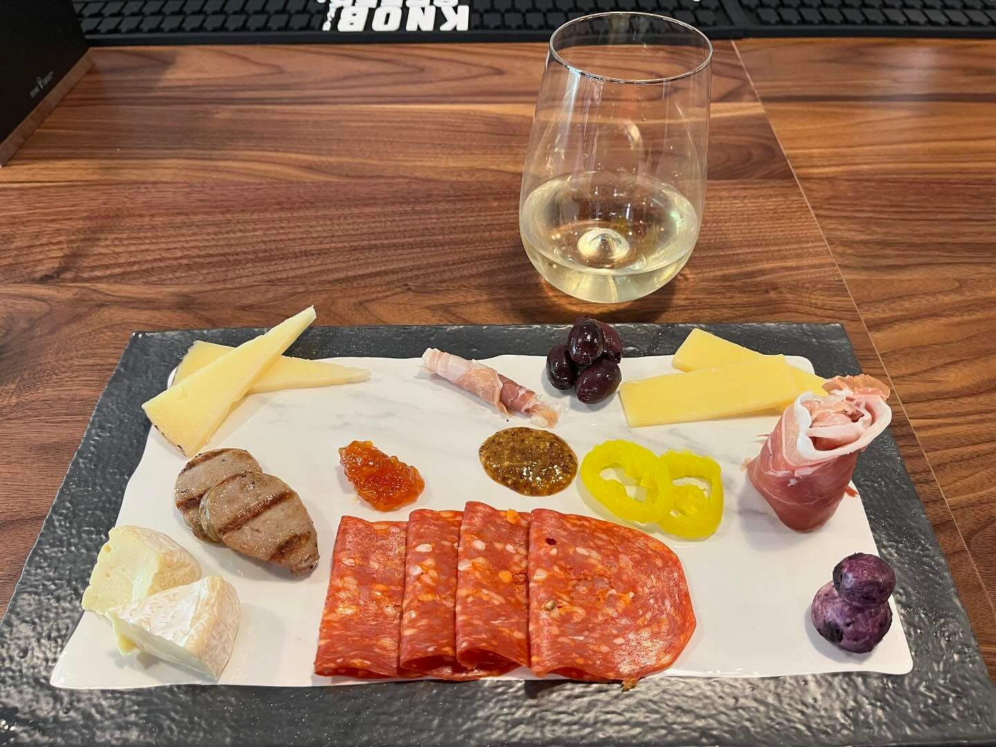 Charcuterie board with cheese, meats, olives, and mustard; a glass of white wine sits nearby on a wood table.
