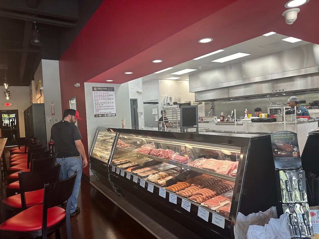 Inside a butcher shop, a customer browses a display case of meats. Red chairs line a counter.