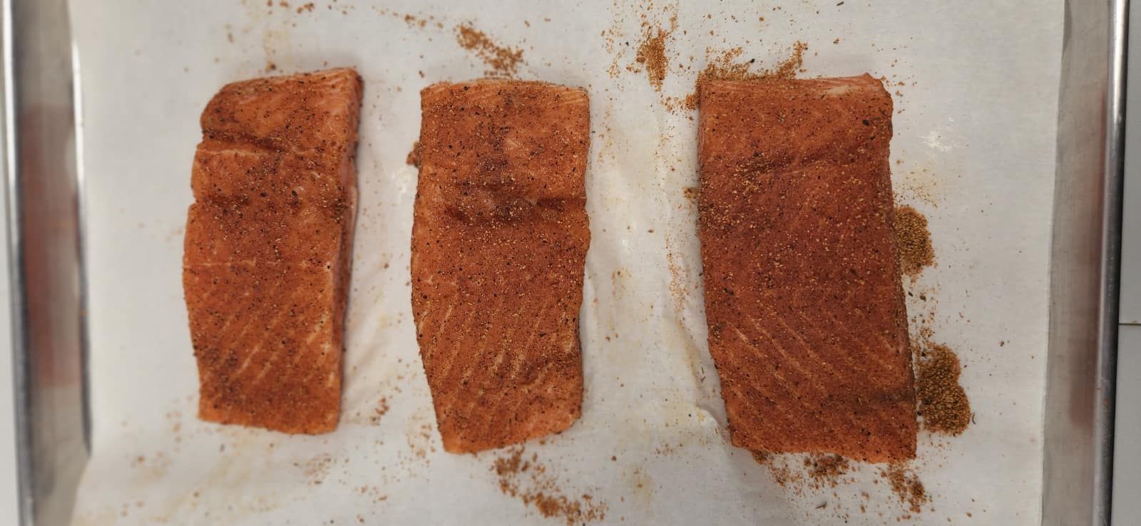 Three seasoned salmon fillets on parchment paper in a baking pan.