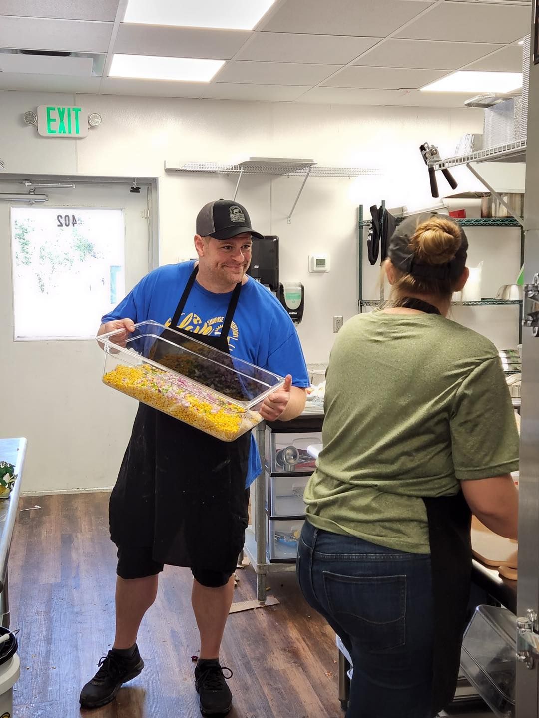 Man holding a container of food smiles at woman in a kitchen.