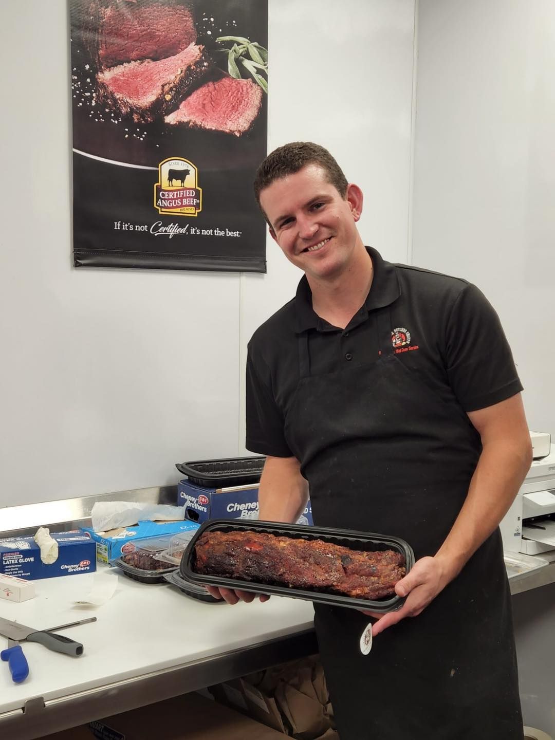 Man smiling, holding a tray of cooked ribs in a commercial kitchen. Advertisement in background.