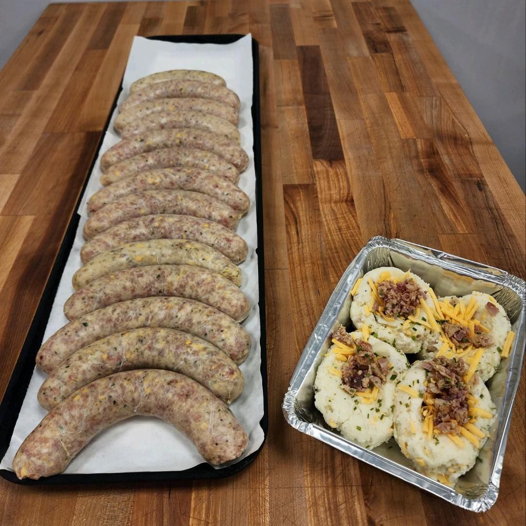 Sausages in a tray next to potato dishes in a foil tray on a wooden surface.
