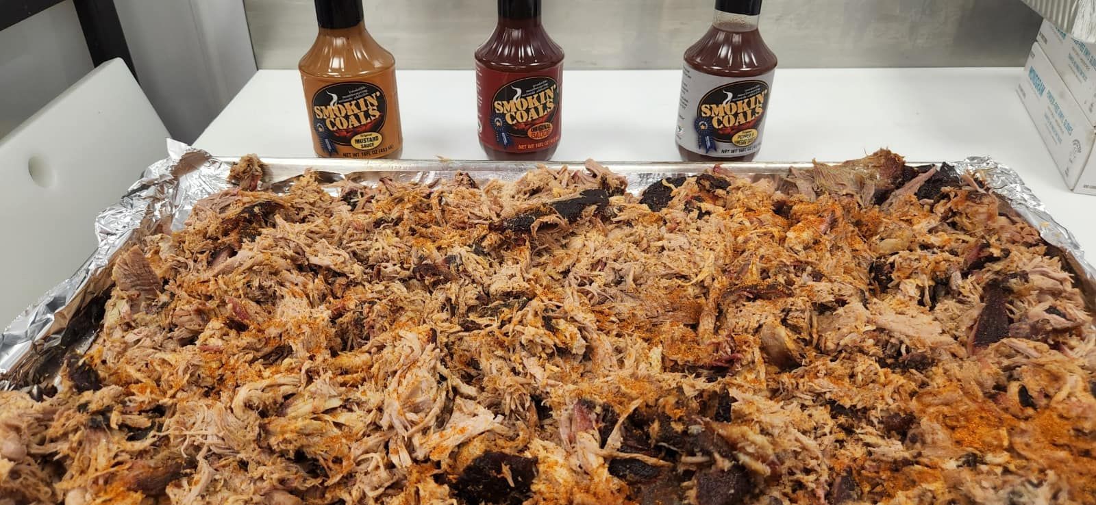 Shredded pork in foil tray with three bbq sauce bottles.