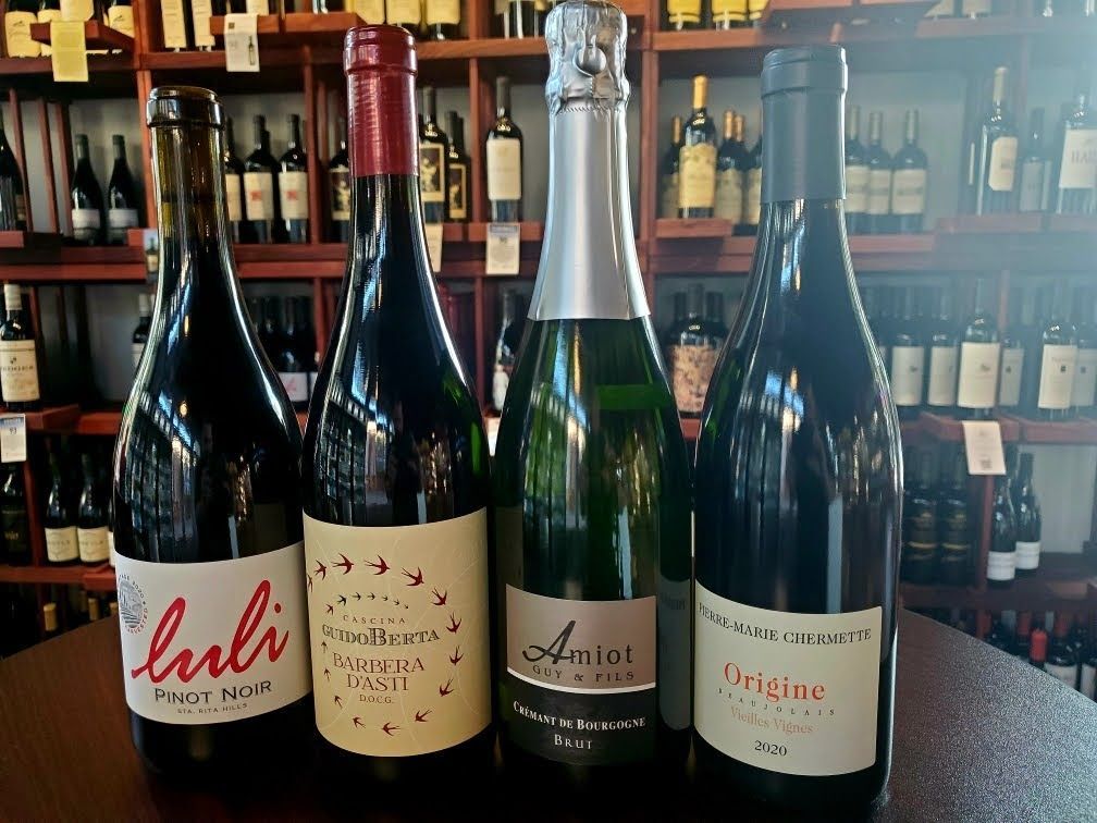 Four wine bottles on display: red, sparkling, and white, in a wine shop setting with bottles on shelves.