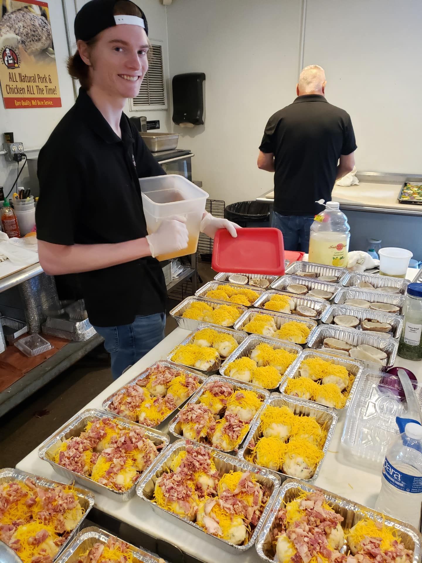 A young man adding liquid to trays of loaded baked potatoes with another person in the background.