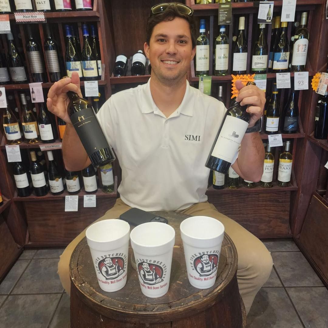 Man holding wine bottles smiles, sitting on a barrel with three cups in a wine store.