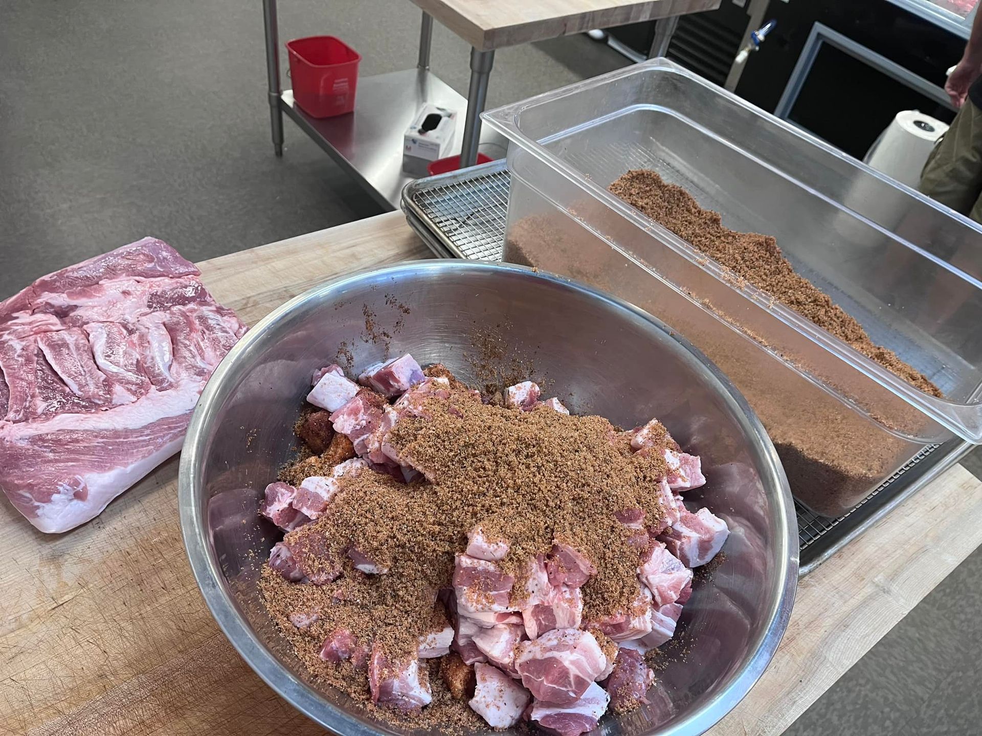 Pork pieces in bowl with seasoning, next to a whole pork cut and container of seasoning, likely for cooking.