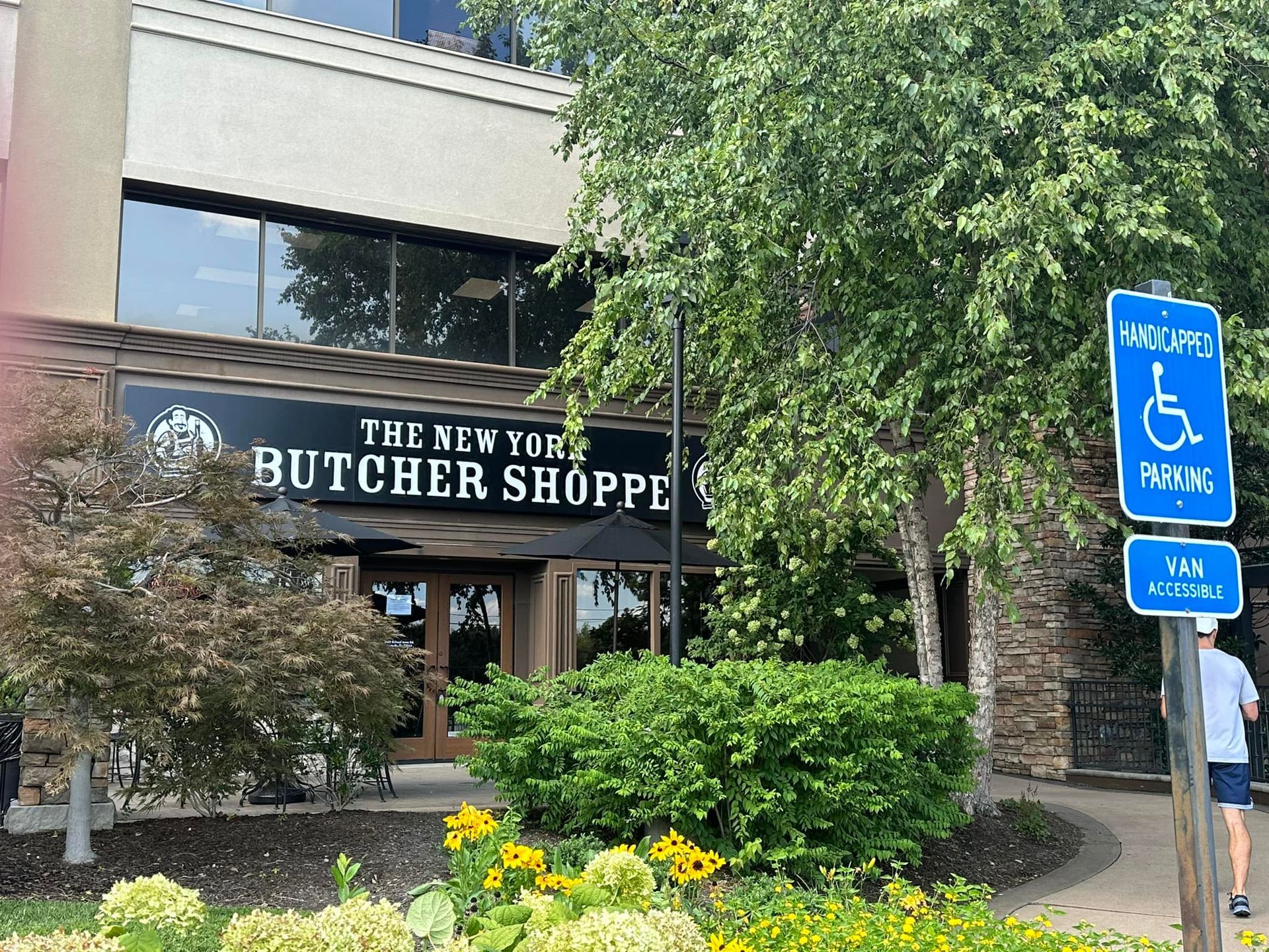 The New York Butcher Shoppe storefront. Handicap parking sign, trees, and a person.