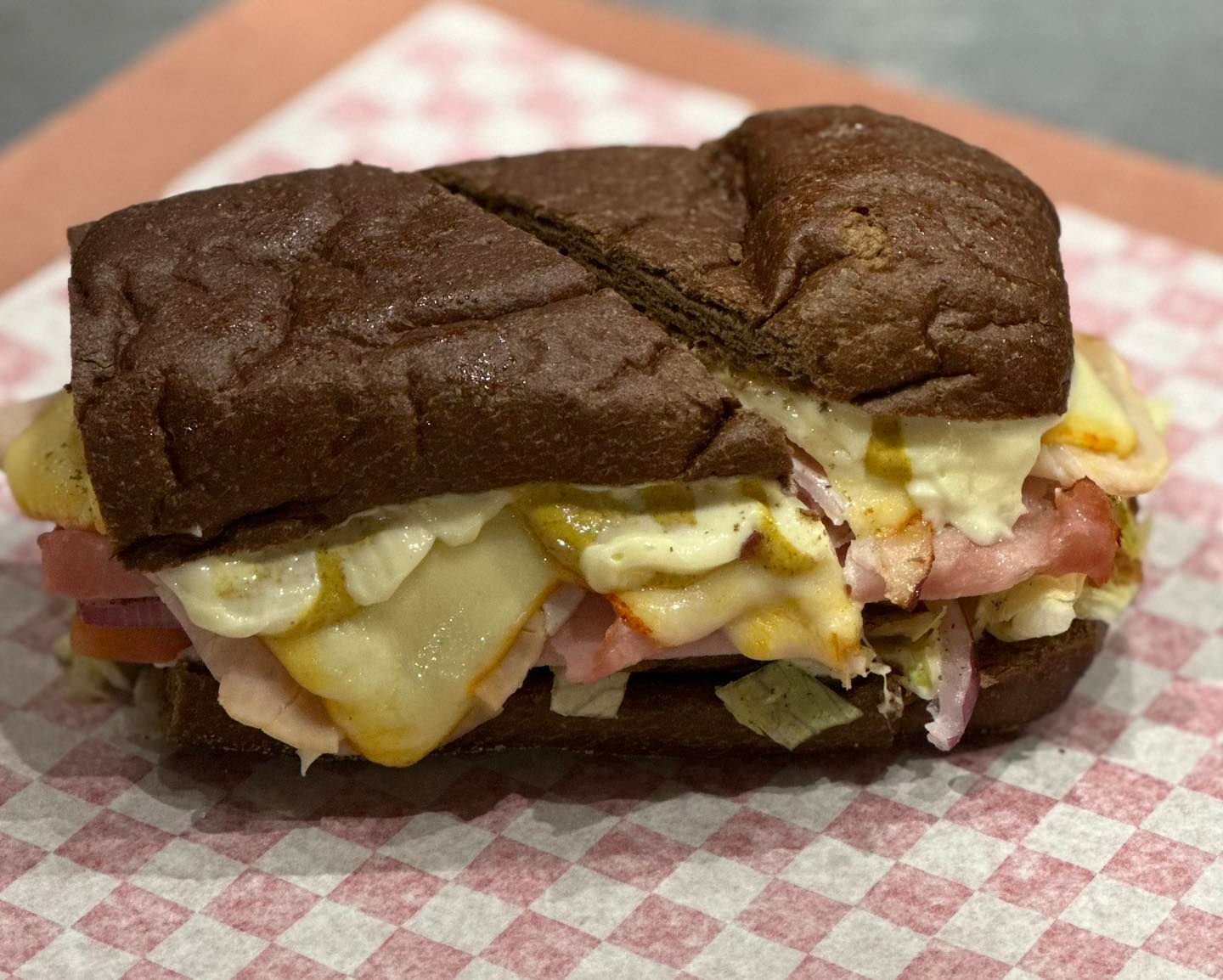 A loaded sandwich on dark bread, with melted cheese, ham, and mustard, on a checkered surface