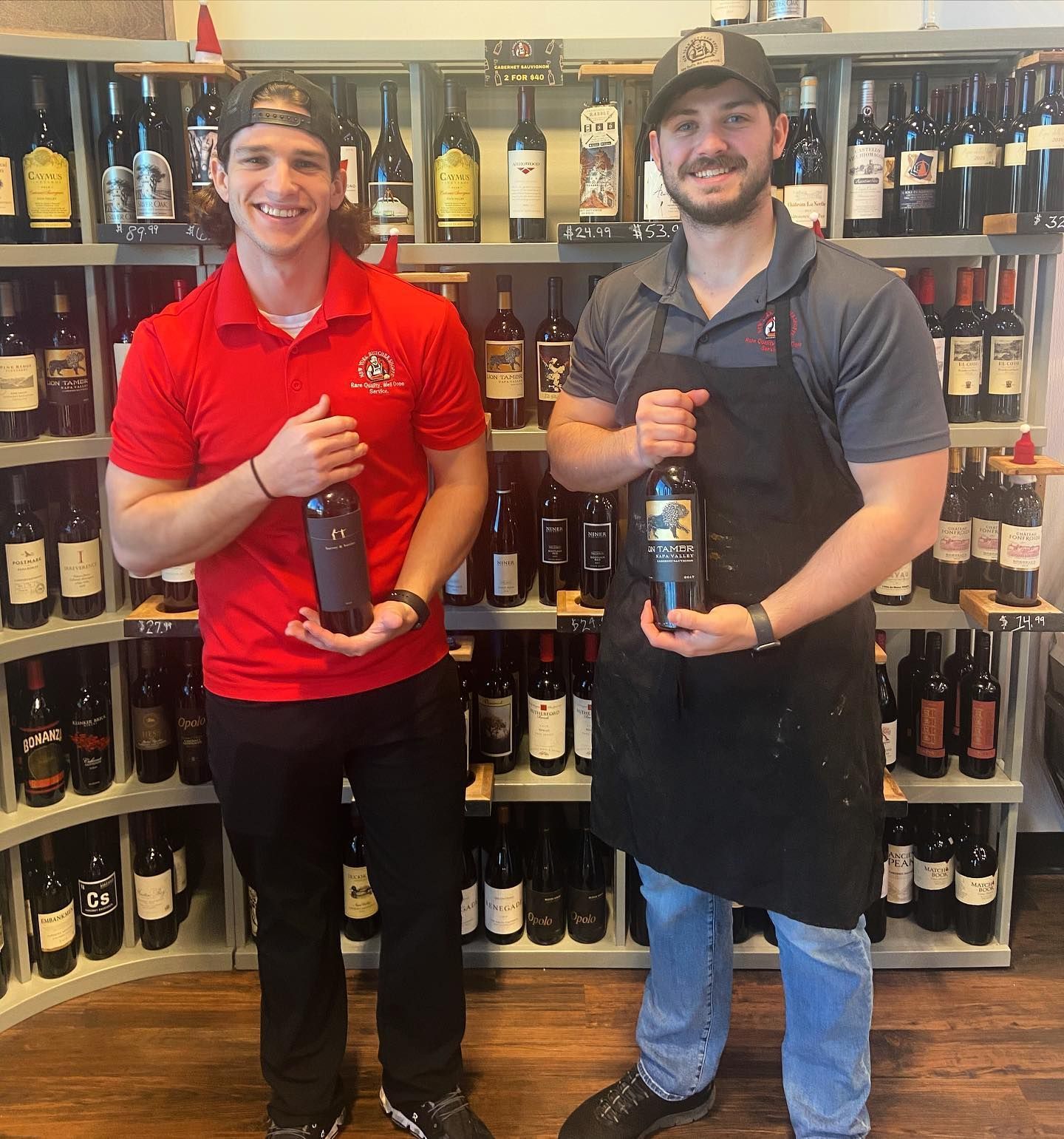 Two men in a wine shop holding bottles, smiling. One in red, one in a black apron, surrounded by shelves of wine.