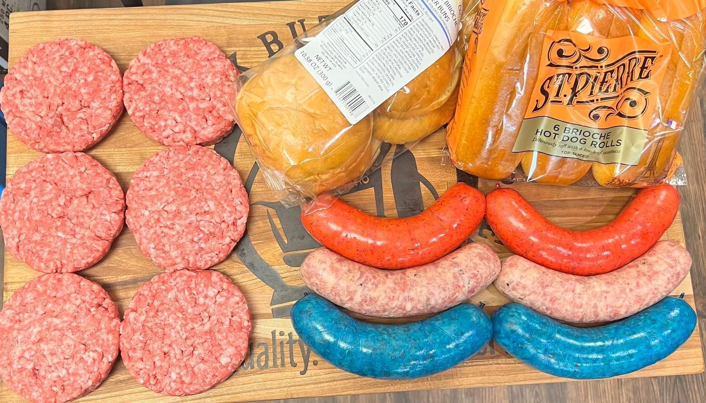 Raw hamburger patties, buns, carrots, and sausages (red, pink, and blue) on a wooden board.