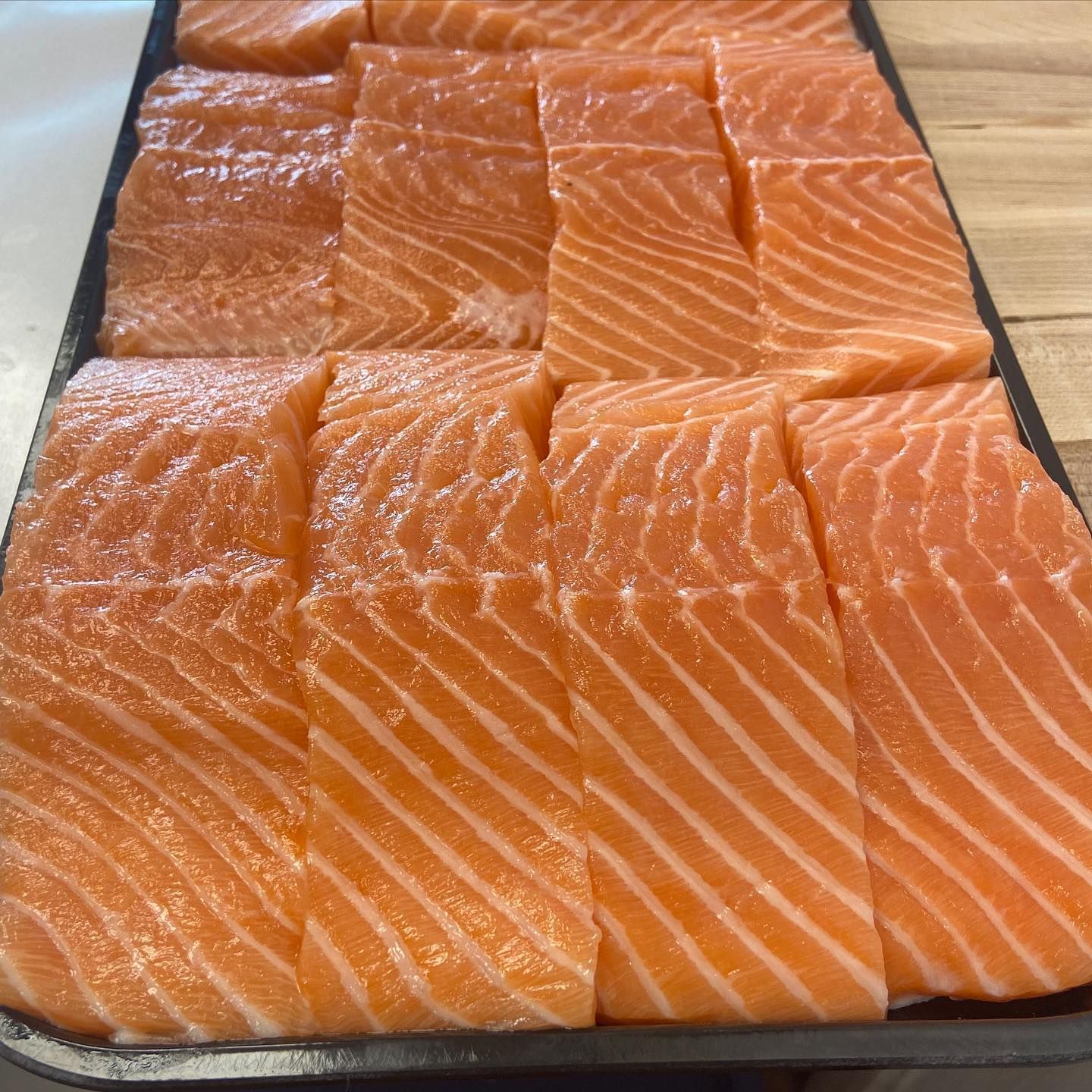 Fresh salmon fillets arranged on a black tray. Orange-pink color, visible marbling.