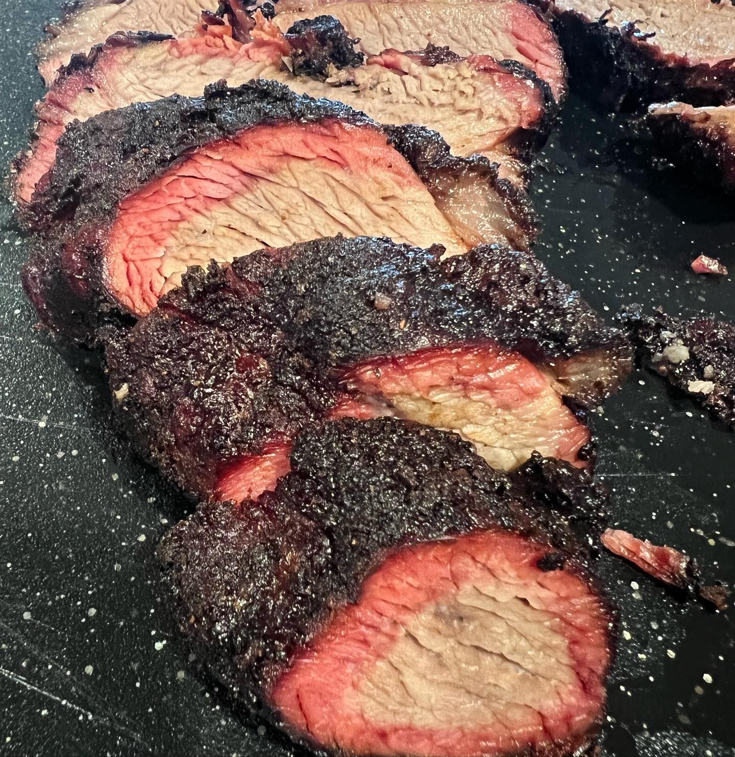 Sliced smoked brisket with a reddish smoke ring and dark bark, on a black surface.