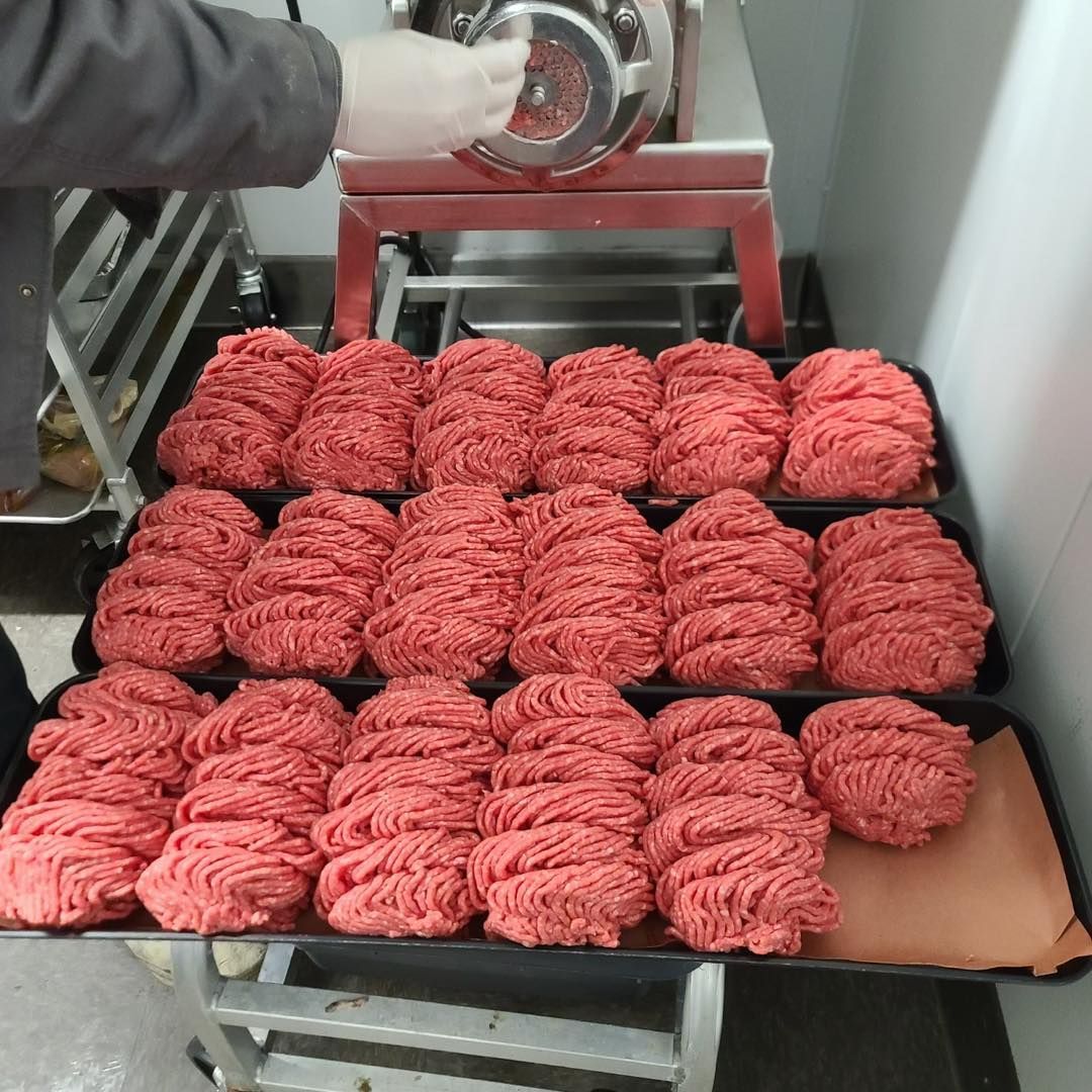 Rows of ground meat portions arranged on trays. A person operates a meat grinder in a commercial kitchen.