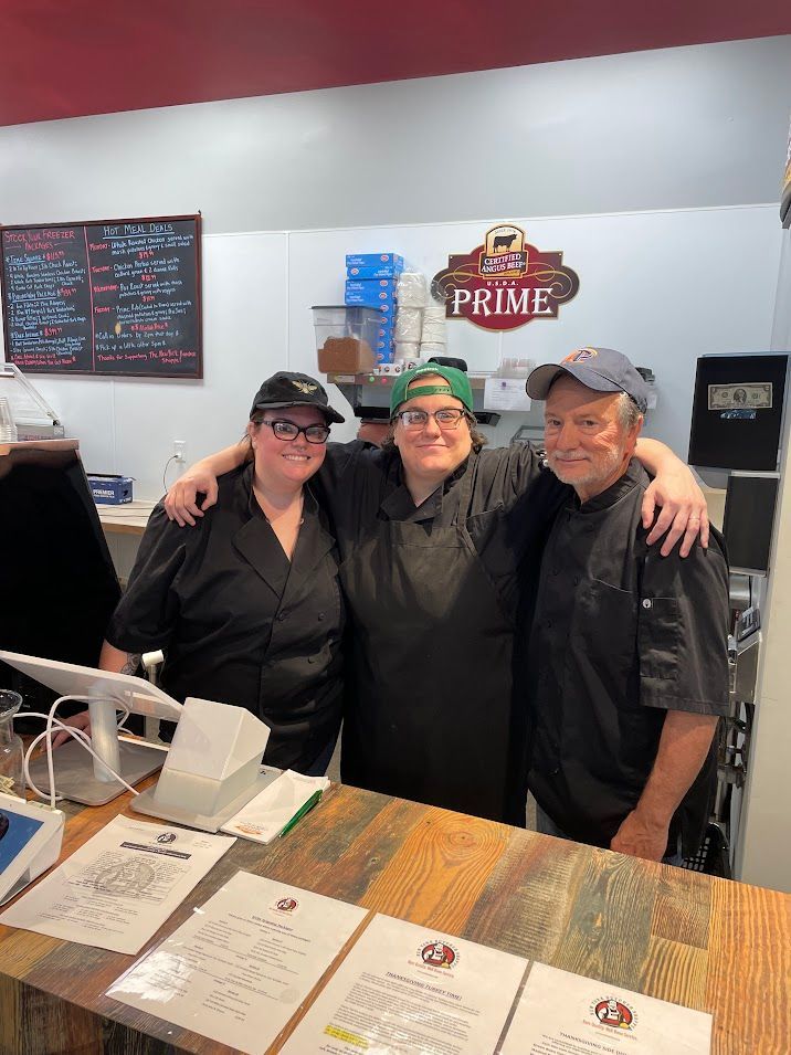 Three restaurant workers smiling behind a counter. Two are in black shirts and hats; one has a green hat.