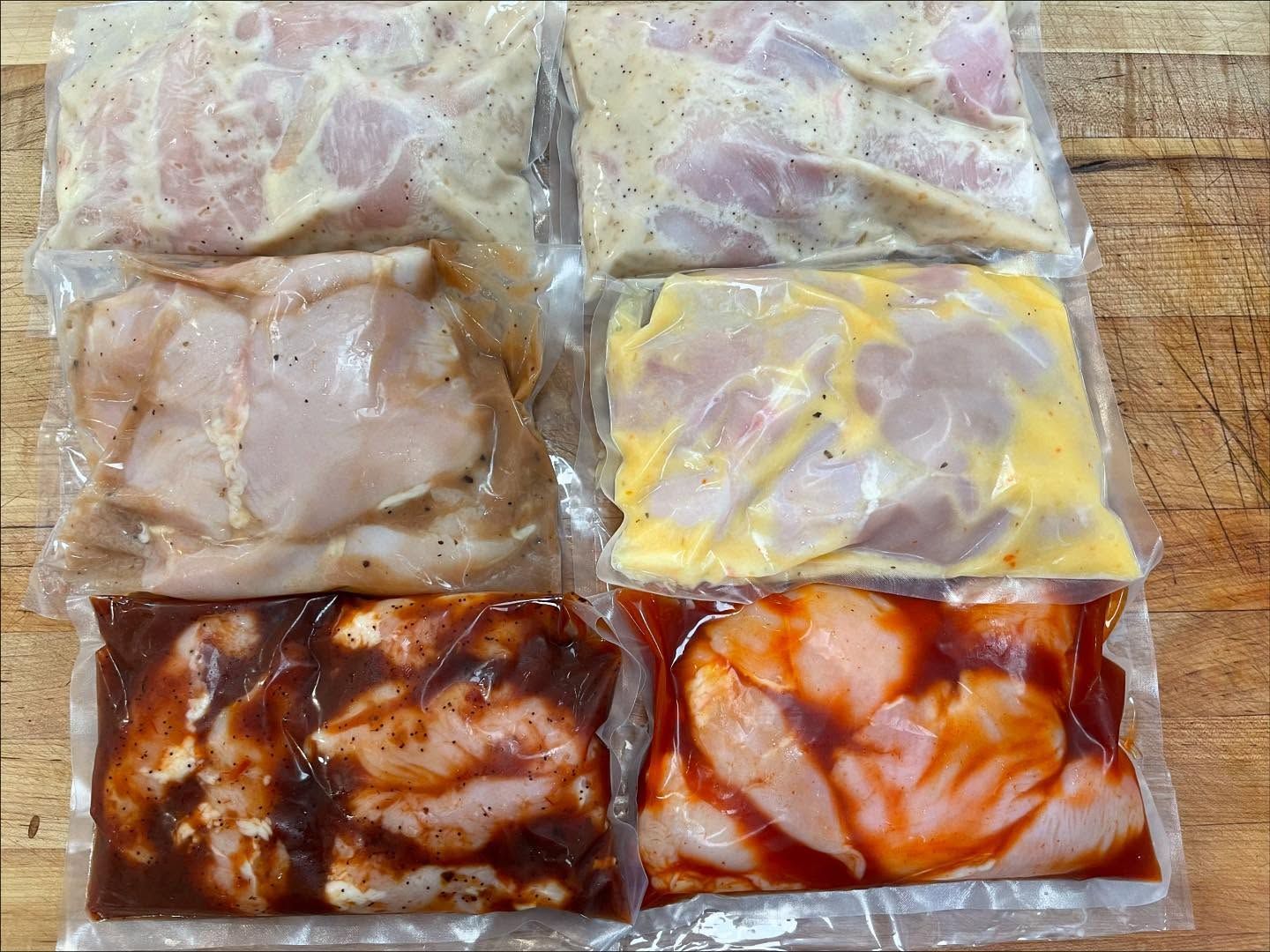 Six sealed bags of marinated chicken breasts, varying colors, arranged on a wooden surface.