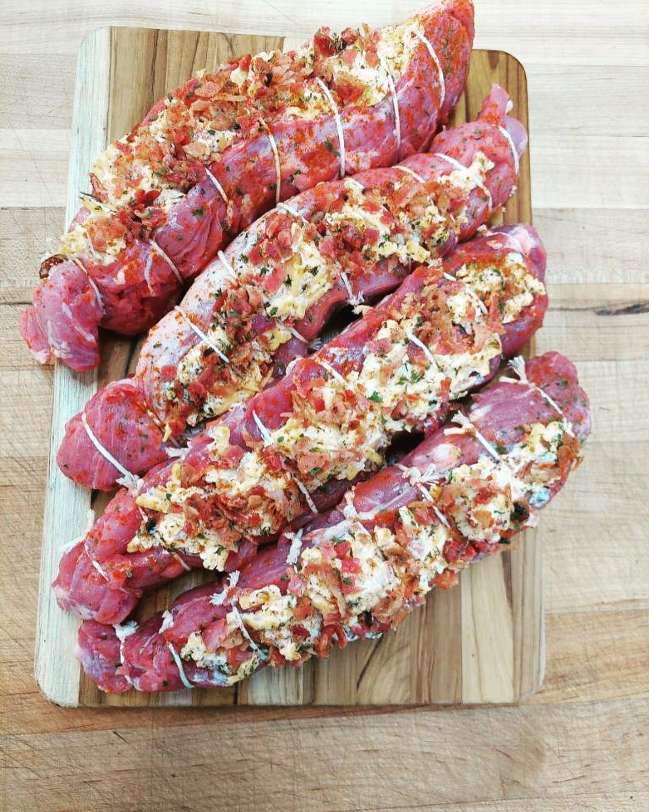 Raw pork tenderloins tied with string, topped with a bacon and cheese mixture, on a wooden cutting board.