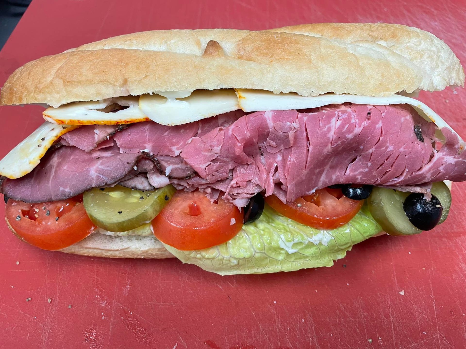Roast beef sandwich on a baguette, layered with cheese, tomato, olives, pickles and lettuce, on red surface.