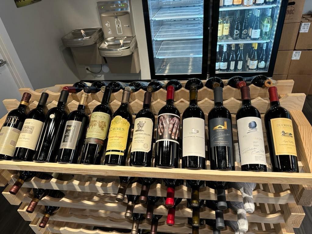 Wine bottles on wooden racks in front of a cooler, indoors.