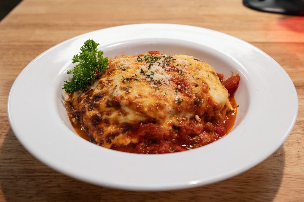Lasagna in a white bowl garnished with parsley and cheese, on a wooden table.