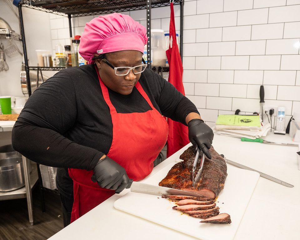 Woman in a red apron and hair covering, slicing brisket with tongs and knife on a cutting board.
