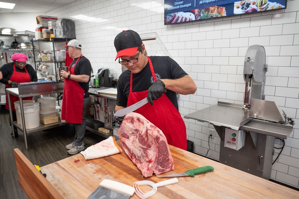 Butcher cutting meat in a butcher shop. Other workers are in the background, the shop is white tiled.