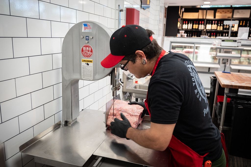 A person in a butcher shop wearing a hat, glasses, and gloves, cutting meat with a band saw.